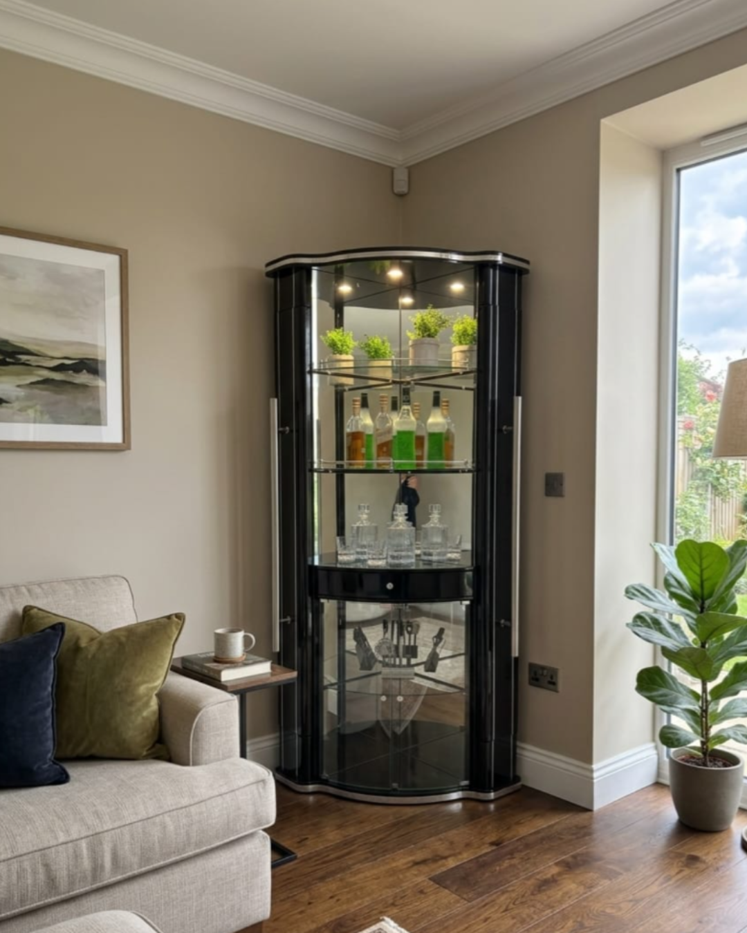 A corner glass display cabinet with a glossy black frame and illuminated shelves, styled in a living room next to a sofa and a large window.