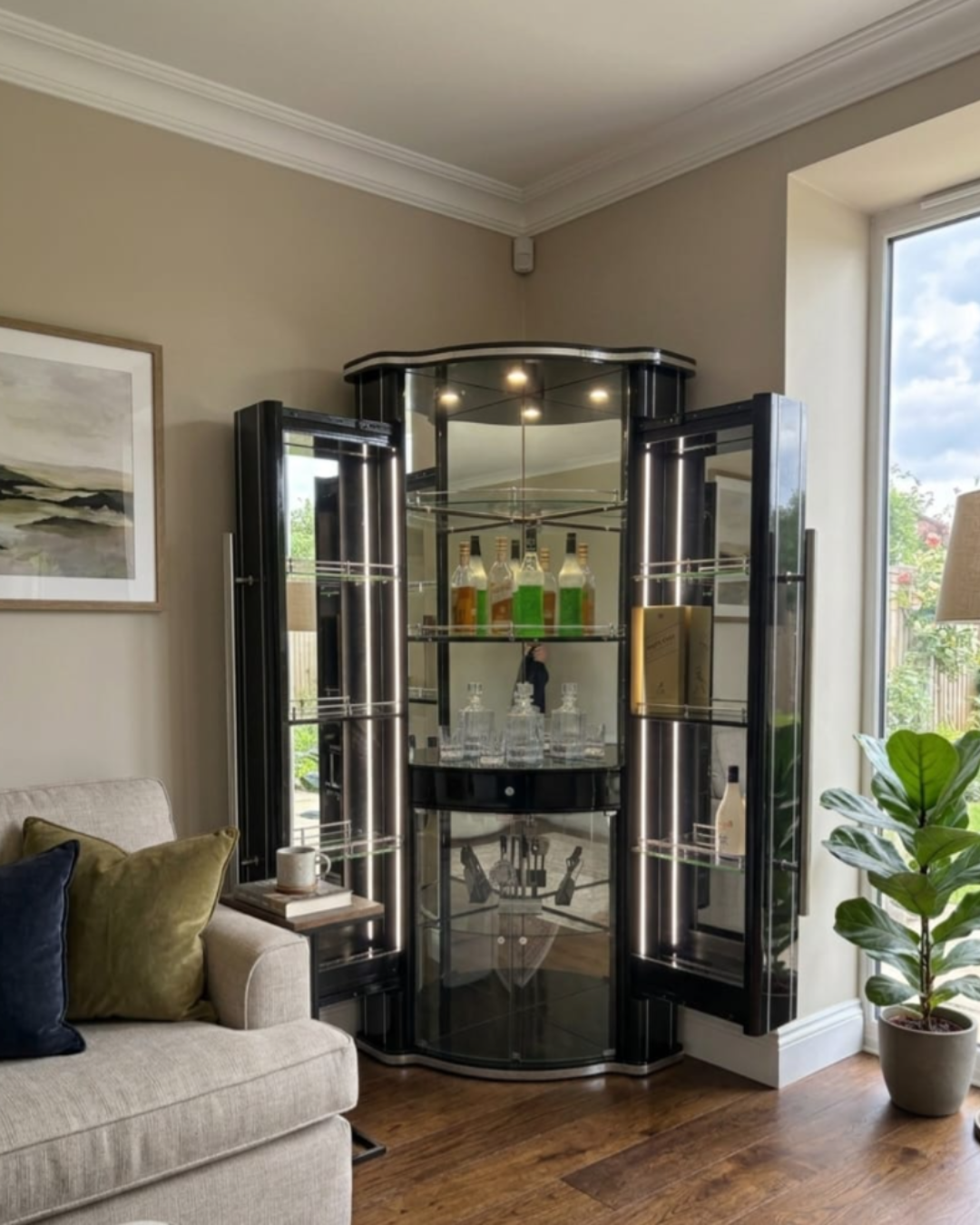 A black corner bar cabinet with glass doors and illuminated shelves, styled in a living room next to a sofa and a potted plant.