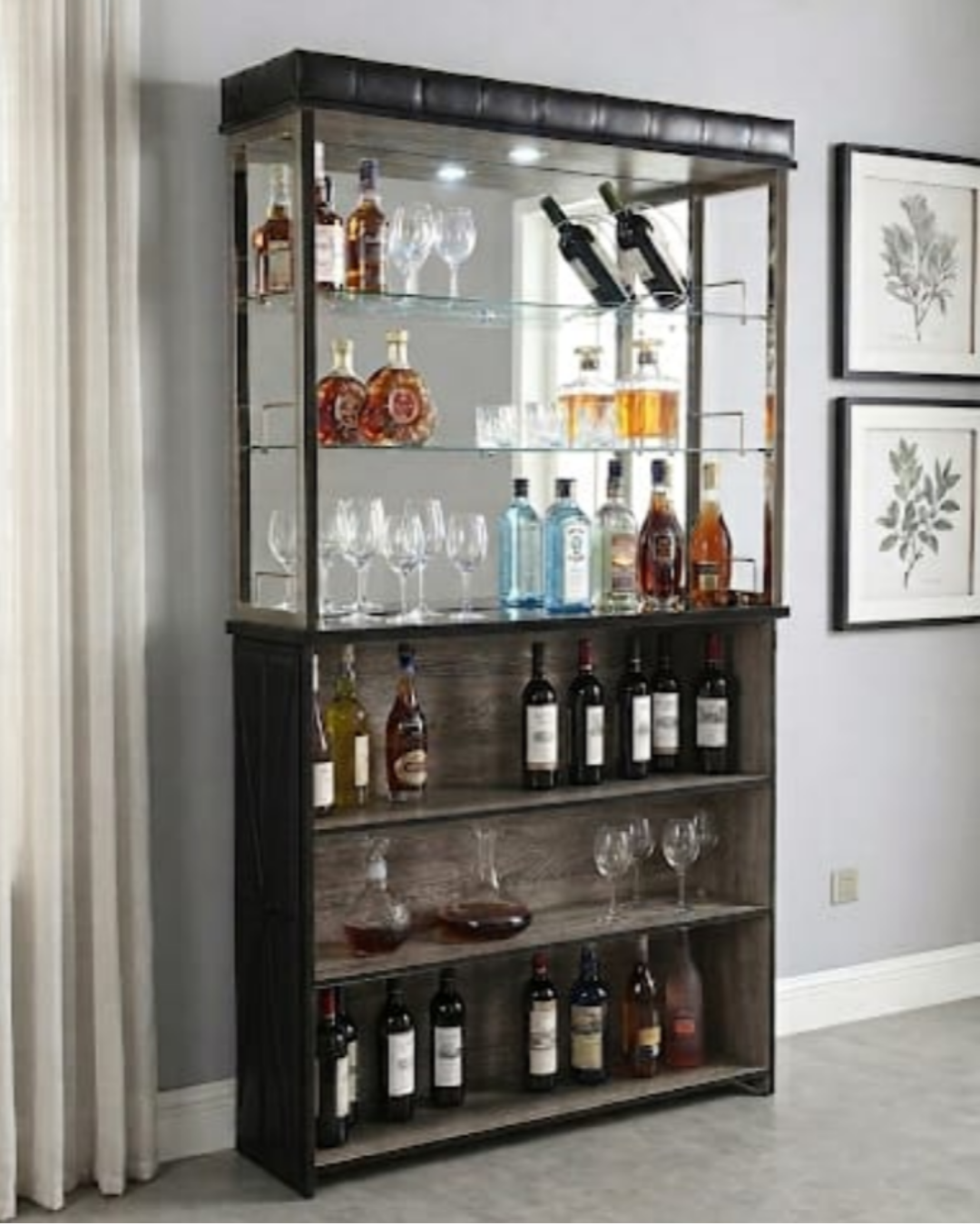 A tall bar cart or wine display cabinet with glass shelves, holding multiple bottles of wine and liquor, and several wine glasses. The cabinet has a dark wood finish with a black top panel and is shown in a home interior setting with a gray wall, framed botanical prints, and a curtain.